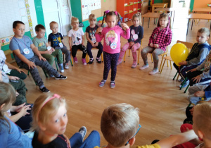 Dzieci siedzą na krzesełkach ustawionych w kole. Bawią się w zabawę integracyjną ,,Podaj balon”. W środku koła stoi dziewczynka, która wykonuje ćwiczenie – wymachy ramion.